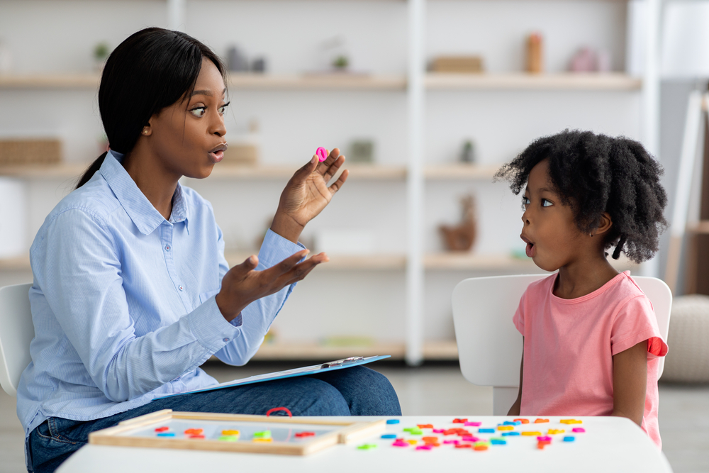 Attractive young african american woman speech therapist working with little black girl, sitting at table with colorful letters, learning pronunciation, clinic interior. Speech disorders in children